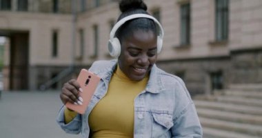 Carefee Afro-Amerikan kadın kulaklık takıyor açık havada müzik dinliyor ve şehirde eğleniyor. Modern yaşam tarzı ve gençlik kavramı.
