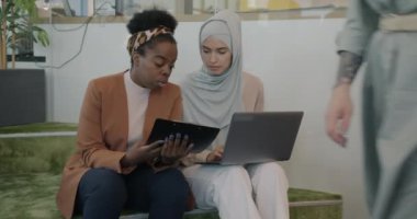 Genç bayan meslektaşlar modern bir iş yerinde dizüstü bilgisayarla konuşuyor ve çalışıyor. İş birliği ve mesleki işgal kavramı.