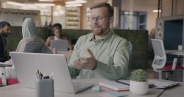 Konuşan ve işaret eden neşeli girişimci açık uzay ofisindeki laptop ve kulaklıkları kullanarak çevrimiçi video görüşmesi yapıyor. İş ve iletişim konsepti.