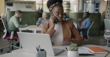 Motive olmuş Afro-Amerikalı iş kadını cep telefonuyla konuşuyor ve açık uzay ofisindeki belgeleri okuyor. İş iletişimi ve danışmanlık istemcisi kavramı.