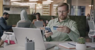 Genç yatırımcı kredi kartı ve akıllı telefonla iş yerinde para transferi yapıyor. Finans ve modern teknoloji kavramı.
