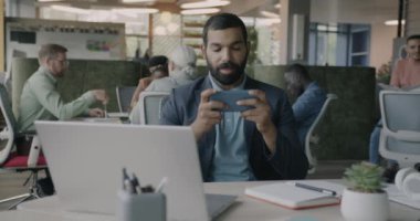 Ofiste akıllı telefonla online video oyunu oynayan neşeli Arap işadamının portresi. Modern teknoloji ve eğlence iş kavramında.