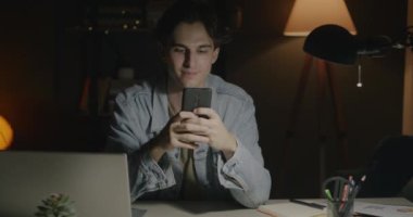 Akıllı telefon kullanan neşeli bir gencin portresi sonra kameraya bakıp geceleri karanlık ofiste gülümseyen. Gadget ve modern yaşam tarzı konsepti.