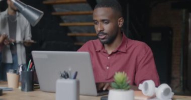 İş arkadaşları arka planda yürürken ve konuşurken dizüstü bilgisayarla çalışan Afrikalı Amerikalı bir adamın portresi. Ofis işleri ve iş adamları kavramı.