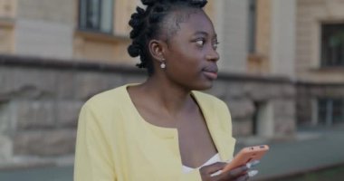 Yakışıklı Afro-Amerikan kadın akıllı telefon kullanıyor ve caddede dikilen şehri izliyor. Modern aletler ve milenyum konsepti.