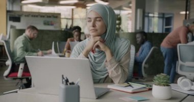 Ofiste masa başında oturan ve kameraya gülen yüzlü Müslüman bir ofis çalışanının yavaş çekimde portresi. İnsan ve iş kavramı.