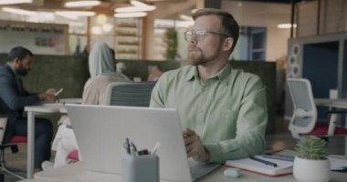 Bilgisayarla çalışan kendine güvenen genç bir adam daktilo yazarken çalışma merkezindeki masada oturan gözlüklere dokunuyor. Modern teknoloji ve çalışan kavramı.