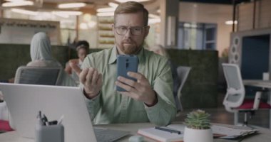 Sosyal medyada müşterilerle iletişim kuran akıllı telefon kullanan genç girişimci portresi ofiste oturuyor. Modern cihazlar ve iş insanları kavramı.