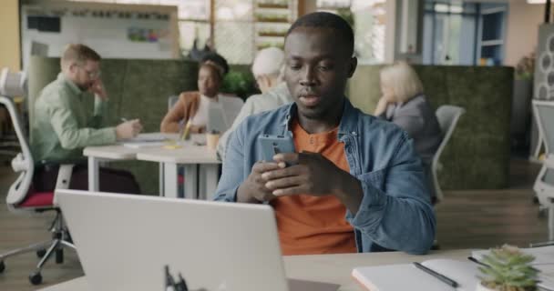 陽気なアフリカ系アメリカ人の従業員は 現代のオフィスでデスクに座ってスマートフォンの笑顔を使用しています インターネット通信と求人コンセプト — ストック動画