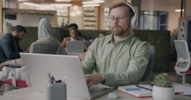 Neşeli genç bir çalışan kulaklıkla müzik dinliyor ve ortak çalışma merkezinde dizüstü bilgisayar kullanıyor. İş adamları ve modern yaşam tarzı kavramı.