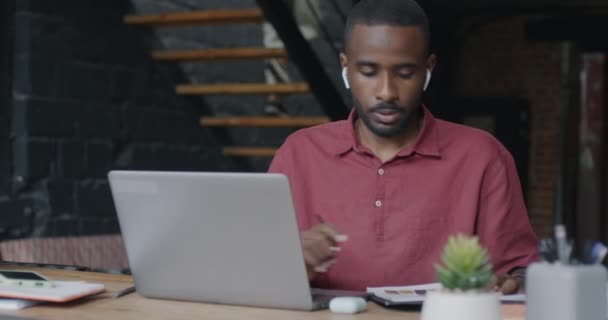 アフリカ系アメリカ人のビジネスマンが コワーキングセンターで仕事を話し合うラップトップとイヤホンを使用してオンラインビデオ通話をする 人とバーチャル会議のコンセプト — ストック動画