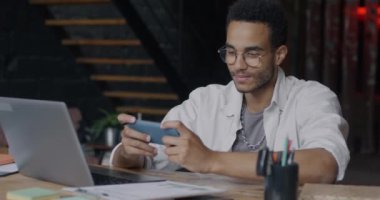 Tasasız Afrikalı Amerikalı bir adam online video oyunu oynuyor ve akıllı telefon yaratıcı ofiste iş yerinde dinleniyor. Milenyum ve eğlence konsepti.