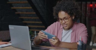 Tasasız Afro-Amerikan iş kadını akıllı telefonuyla online video oyunu oynuyor ve ofisteki uygulamadan keyif alıyor. İş ve eğlence konsepti.