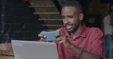 Afrika kökenli Amerikalı iş adamı iş yerinde çalışırken akıllı telefonuyla mobil video oyunu oynuyor. Malzemeler ve eğlence konsepti.