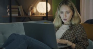 Dizüstü bilgisayarla çalışan serbest çalışan genç bir kadın geceleri evdeki kanepede oturuyor. Modern teknoloji ve serbest meslek kavramı.