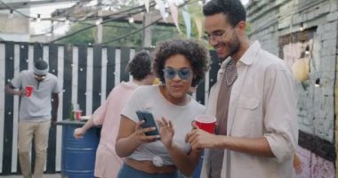 Afro-Amerikalı erkek ve kadın arkadaşlar konuşuyor ve akıllı telefon kullanıyor. Kafedeki açık hava partisinde ekrana bakıp gülüyorlar. Gadget ve modern yaşam tarzı konsepti.