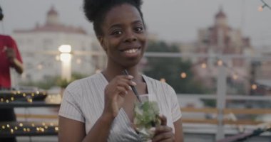 Geceleri çatıda elinde kokteyl bardağıyla duran muhteşem Afrikalı Amerikalı kadının yavaş çekimde portresi. Neşeli DJ müziği ve dansı arka planda karıştırıyor.