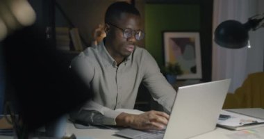 Afro-Amerikalı iş adamı dizüstü bilgisayarını kullanıyor ve akşam ofisteki not defterine bilgi yazıyor. Profesyonel meslek ve e-ticaret kavramı.