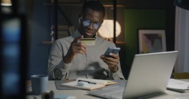 Akıllı telefon kullanan Afrikalı Amerikalı bir adam kredi kartı alışverişi yapıyor ve geceleri evde ödeme yapıyor. Finans ve ticaret kavramı.