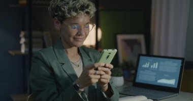 Genç bir kadın akşamları evden çalışırken modern akıllı telefon mesajları kullanıyor. Çağdaş teknoloji ve hırslı iş insanları kavramı.