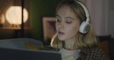 Kulaklıkla müzik dinleyen, şarkıdan keyif alan ve akşam evde dizüstü bilgisayar kullanan mutlu genç bir kadın. Milenyum ve teknoloji kavramı.