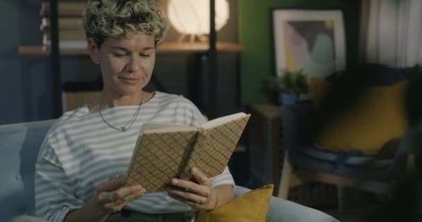Веселая Молодая Женщина Читает Книгу Улыбаясь Расслабляясь Уютной Квартире Вечером — стоковое видео