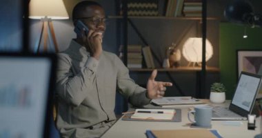 Afrika kökenli Amerikalı ofis çalışanı cep telefonuyla konuşuyor ve akşamları karanlık ofiste çalışırken notlar alıyor. İş iletişimi ve iş kavramı.