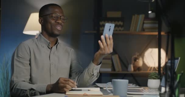 アフリカ系アメリカ人の起業家で 夜の闇のオフィスでスマートフォンメッセンジャーを使用してオンラインビデオ通話をしています 人とプロフェッショナルなコミュニケーションのコンセプト — ストック動画