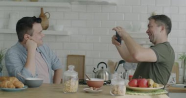 Erkek arkadaşı mutfak masasında akıllı telefonuyla fotoğraf çekerken kameraya poz veren neşeli eşcinsel adam. Fotoğraf ve aynı cinsiyet ilişkisi kavramı.