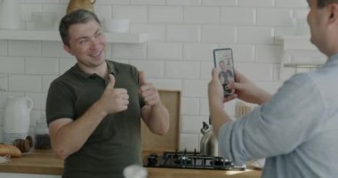 Sevgi dolu koca mutfakta akıllı telefonuyla fotoğraf çekerken tasasız eşcinsel adam iyi niyet göstergesi olarak poz veriyor. Mutlu çift dairede eğleniyor..