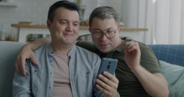 Akıllı telefon kullanan neşeli eşcinsel çift evdeki kanepede oturup sosyal medyanın tadını çıkarıyor. Modern teknoloji ve LGBTQ ilişki kavramı.