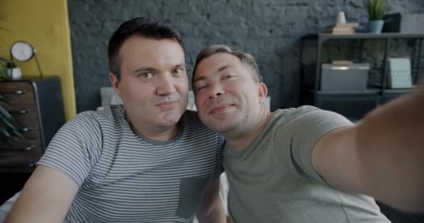 这对可爱的同性恋夫妇在镜头前摆出笑脸 自拍坐在床上的肖像 男人在镜头前玩的开心 — 图库视频影像