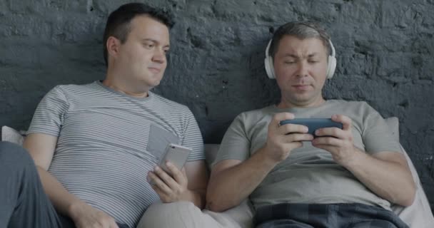 ベッドに横たわって見ている彼氏の間 スマートフォンでビデオゲームをするヘッドフォンを身に着けている楽しい男 モダンテクノロジーとゲイカップルのライフスタイルコンセプト — ストック動画