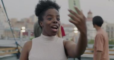 Mutlu Afro-Amerikalı kadın akıllı telefonuyla çevrimiçi video görüşmesi yapıyor. Çatıdaki partide dans eden insanları işaret ediyor. İletişim ve modern aygıtlar kavramı.