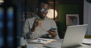 Afrika kökenli Amerikalı işadamı, gece karanlığında akıllı telefon ve kredi kartı kullanıyor. Bankacılık ve finans kavramı.