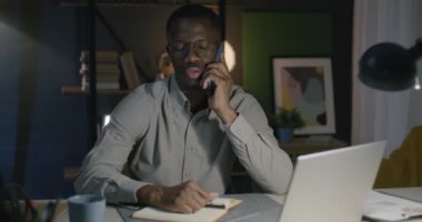 Cep telefonuyla konuşan ve çay içen Afro-Amerikalı bir adam gece masa başında oturuyor. Profesyonel iletişim ve insan konsepti.