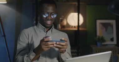 Afrikalı Amerikalı iş adamı akıllı telefonuyla online video oyunu oynuyor. Geceleri ofiste eğleniyor. İş adamı ve oyun konsepti.