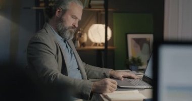 Not defterine bilgi yazan orta yaşlı bir iş adamı geceleri ofiste çalışan dizüstü bilgisayarlara bakıyor. İş ve iş kavramı.