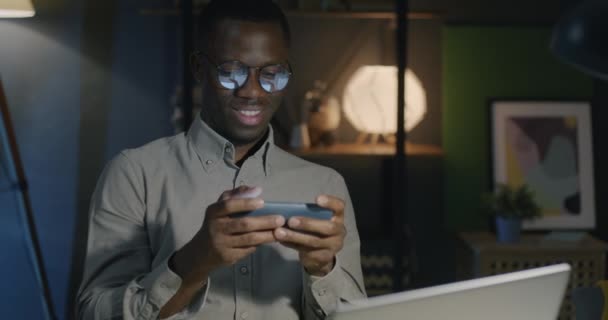 アフリカ系アメリカ人のビジネスマンは 夜にオフィスで楽しんでいるスマートフォンでオンラインビデオゲームをプレイします ビジネスマンとゲームコンセプト — ストック動画