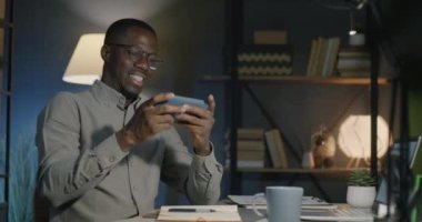 Kaygısız Afrikalı Amerikalı adam, akıllı telefonuyla online video oyunu oynuyor ve geceleri karanlık ofiste mobil uygulamanın tadını çıkarıyor. Oyun ve işyeri kavramı.