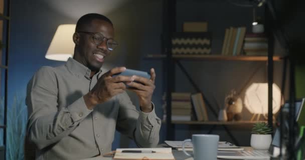 夜のダークオフィスでモバイルアプリを楽しんでいるスマートフォンでオンラインビデオゲームをプレイするケアフリーのアフリカ系アメリカ人男性 ゲームと職場のコンセプト — ストック動画