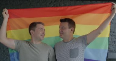İçerideki kameraya gülümseyen gökkuşağı LGBTQ bayrağı tutan mutlu eşcinsel çiftin yavaş çekim portresi. Cinsiyet eşitliği ve aynı cinsiyet ilişkisi kavramı.