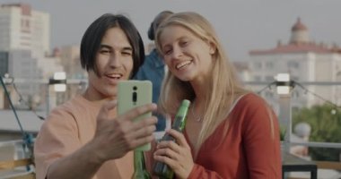 Asyalı bir adam ve beyaz bir kadın akıllı telefon kamerası için poz veriyor çatıdaki partide selfie çekiyorlar. Modern teknoloji ve eğlence konsepti.
