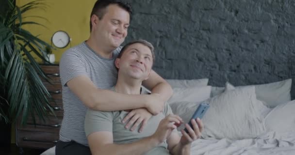 爱同性恋夫妇坐在床上拥抱 用智能手机在家中享受浪漫时光的慢动作 小武器和同性恋关系概念 — 图库视频影像