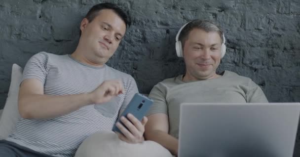 自宅でヘッドフォンで音楽を聞くラップトップとスマートフォンを使用してベッドに横たわるケアフリーのゲイ男性 モダンデバイスとLgbtqコンセプト — ストック動画