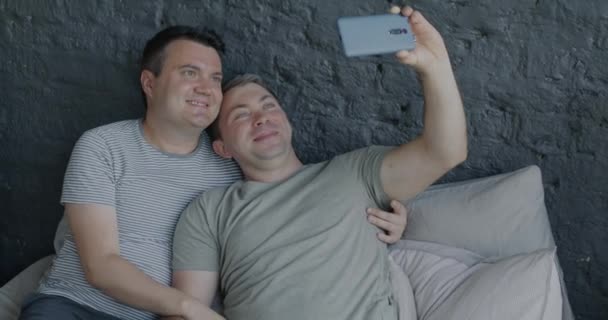 快乐的同性恋男人摆出一副智能手机相机的姿势 自拍躺在床上拥抱 享受家里的闲暇时光 照片和男女同性恋 双性恋和变性者关系概念 — 图库视频影像