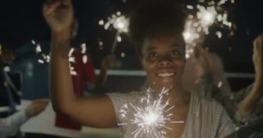 Geceleri bengal ışığıyla dans eden çekici Afrikalı Amerikalı kadının yavaş çekim portresi. Kameraya bakarak arkadaşlarıyla eğleniyor.