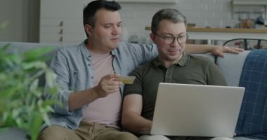 İnternetten alışveriş yapan ve dairede dizüstü bilgisayarla ödeme yapan iki yakışıklı gay çift. Bankacılık ve tüketim kavramı.