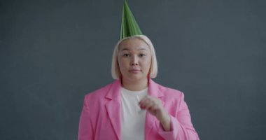 Mutsuz ve yalnız Asyalı bayanın ağır çekimde portresi parti şapkası takan, ıslık çalan ve iç çeken gri arka planda hüznü ifade eden.