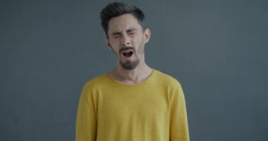 Gri arka plandaki kameraya bakarak esneyen, esneyen genç bir adamın yavaş çekim portresi. İnsanlar ve yüz ifadesi kavramı.